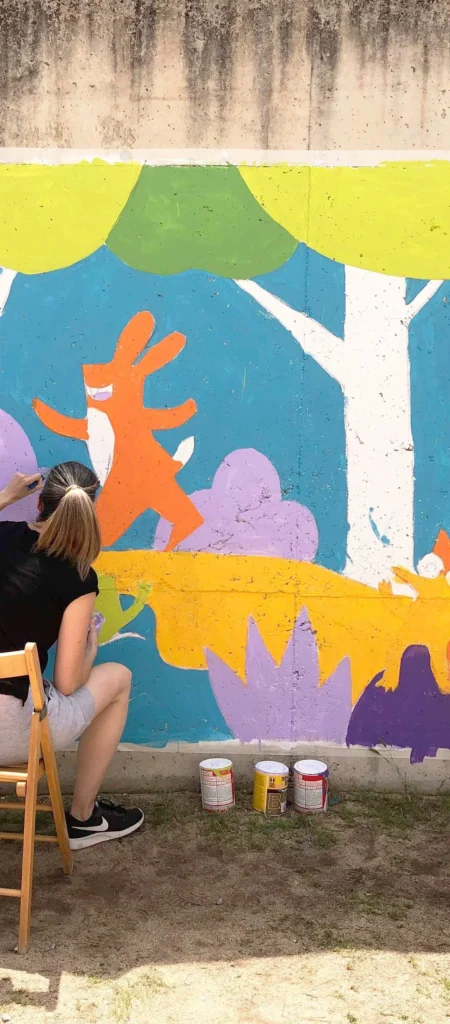 Mireia Serra asseguda en una cadira pintant a mà alçada un mural infantil sobre un mur de formigó amb un conill taronja i elements vegetals en verd, lila i groc